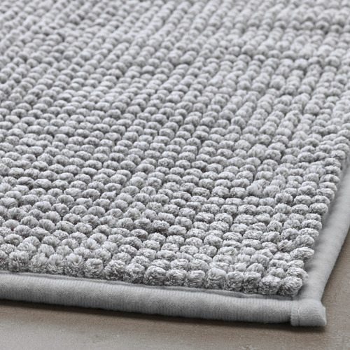 Alfombra de baño suave de color gris claro con superficie texturizada, plegada sobre suelo de madera.