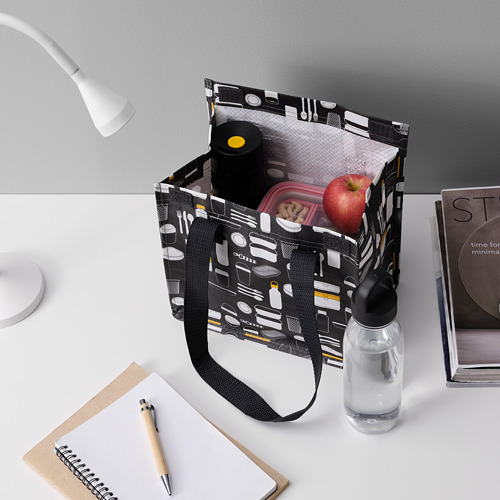 Bolsa de almuerzo negra con asas que contiene una botella, una manzana y un recipiente, colocada sobre un escritorio blanco con un cuaderno, un bolígrafo y una lámpara.