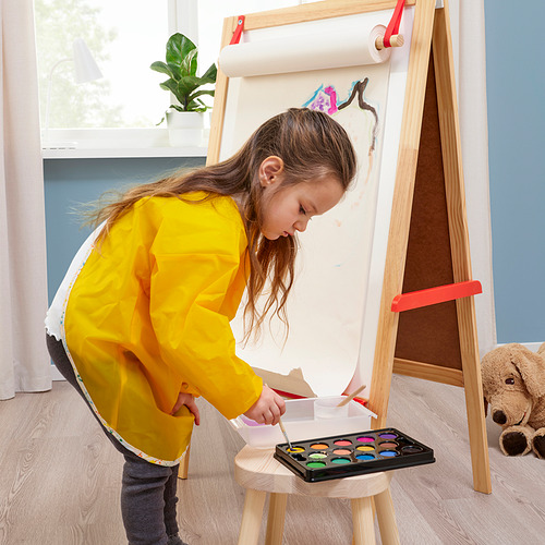 Un niño con un delantal amarillo está de pie frente a un caballete de madera, pintando con acuarelas.