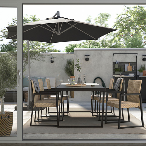 Moderno patio al aire libre con mesa de metal negro, sillas de mimbre y una gran sombrilla negra. Paredes grises, plantas y una zona de cocina al fondo.