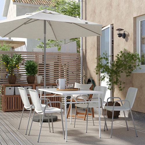 Moderno juego de comedor al aire libre con sillas y mesa blancas, gran sombrilla beige, plantas en macetas, cercas de madera y edificio beige en el fondo.
