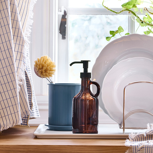 Junto a la ventana, el mostrador de madera sostiene un cepillo para platos en taza azul, dispensador de jabón y platos apilados en el soporte. Las cortinas cuelgan al lado de la ventana.