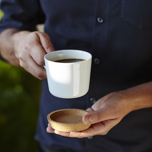 Persona sosteniendo una taza de cerámica blanca con líquido oscuro y un pequeño posavasos, con IKEA 365+, diseñado para proteger las superficies de la mesa.