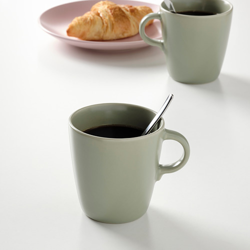 Dos tazas verdes con cucharas, una llena de café, junto a un plato con un croissant.