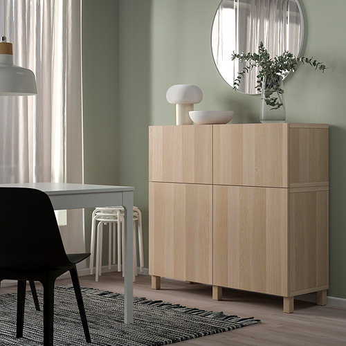 Armario BESTÅ de madera clara con puertas en un salón moderno. Cubierto con jarrones y vegetación. Es alto y estrecho, y cabe bien bajo un espejo redondo.