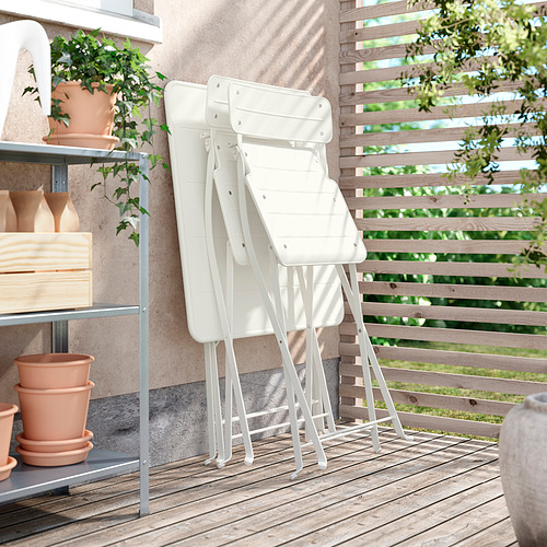 Dos sillas plegables de metal blanco apiladas al aire libre sobre una terraza de madera cerca de una maceta de plantas y estanterías con elementos de decoración, ideales para espacios pequeños.