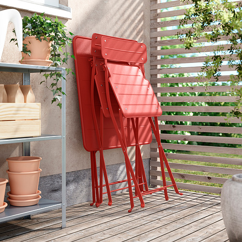 Tres sillas de metal rojo apiladas, colocadas al aire libre sobre una cubierta de madera. Rodeado de plantas en macetas y una estantería con más macetas.