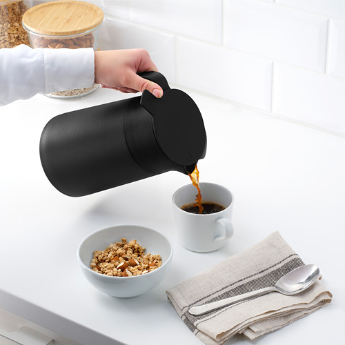La persona vierte el café de un matraz en una taza junto a la granola, la cuchara y las servilletas.