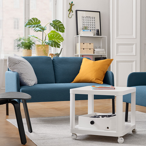 Sala de estar moderna con sofá azul, almohada amarilla, mesa de centro blanca, plantas y decoración.