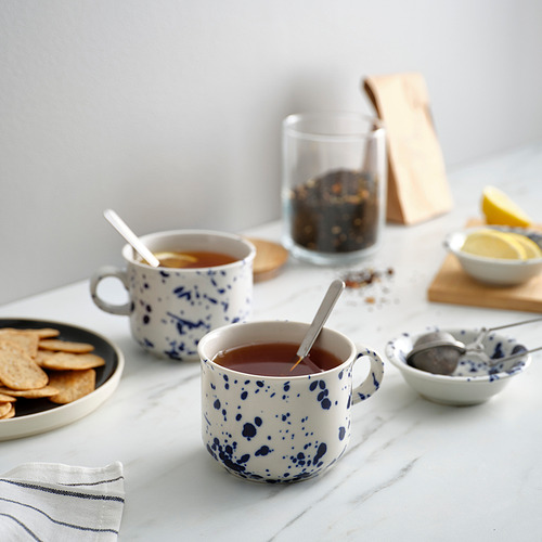 Dos tazas blancas SILVERSIDA con puntos azules. Uno sostiene té, cuchara adentro. Más pequeño que los platos con galletas saladas y rodajas de limón. Fabricado en gres.