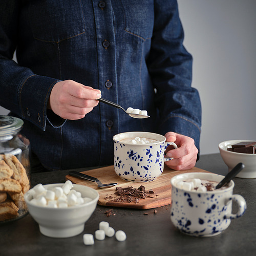 Persona que agrega malvaviscos a una taza sobre una tabla de madera. La taza es blanca con puntos azules, rodeada de malvaviscos, galletas y una cuchara.