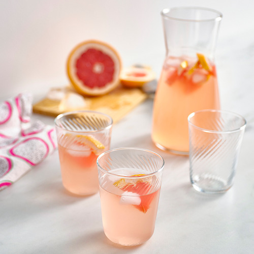 Juego de vasos KLIPPLAX: tres bebidas rosas en vasos finos y acanalados junto a una jarra más grande, pomelo cortado por la mitad y servilletas estampadas sobre una tabla de madera.