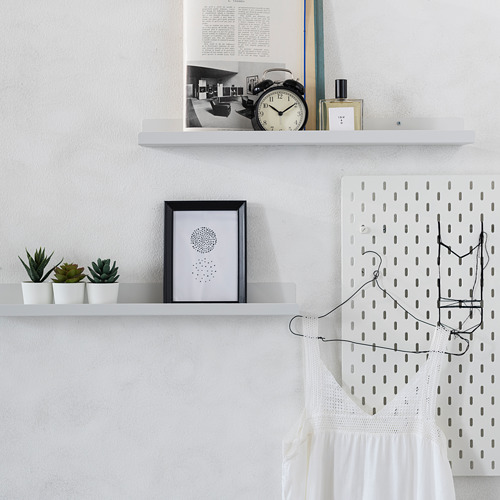 Tres estantes blancos con detalles en negro sostienen artículos como un reloj, plantas y arte. A su lado hay un tablero de clavijas con luces colgantes y un vestido blanco.