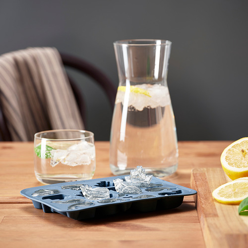 Bandeja de cubitos de hielo con formas juguetonas sobre mesa de madera, junto a un vaso de agua y una jarra, perfecta para las bebidas de verano.