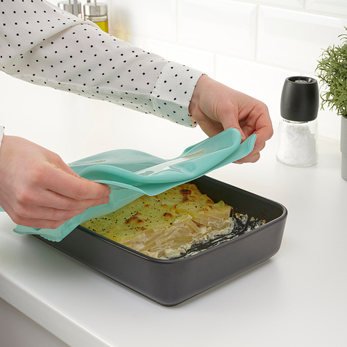 Persona colocando una cubierta de silicona verde en una fuente para hornear con alimentos cocidos, realzando la frescura.