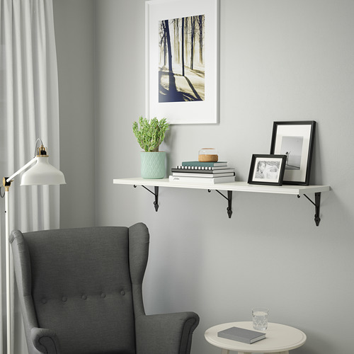 Un rincón acogedor con un sillón gris junto a una estantería de pared blanca que exhibe una planta, libros, marcos y una lámpara.