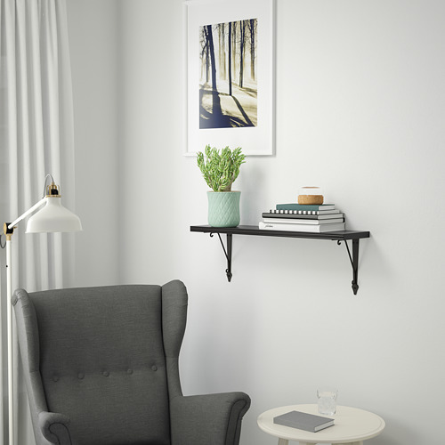 Habitación minimalista con estantería de pared BERGSHULT negra. Exhibiendo libros, plantas y decoración sobre una silla gris. Obra de arte enmarcada en blanco arriba.