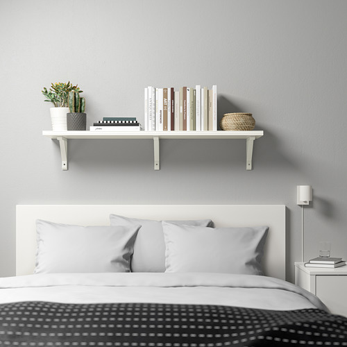 Dormitorio minimalista con estante de pared blanco BERGSHULT que sostiene libros y plantas sobre una cama con almohadas grises y manta negra estampada.