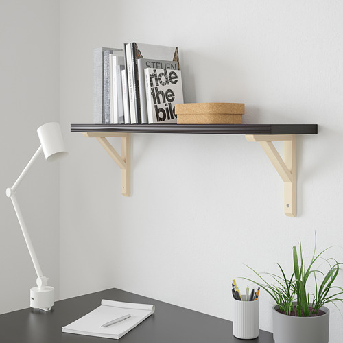 Estantería de pared BERGSHULT negra con soportes de madera que sostiene libros y objetos pequeños junto a un escritorio con una lámpara, un cuaderno, bolígrafos y una planta.