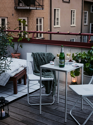 Balcón moderno con mesas y sillas plegables blancas, manta verde, copas de vino, velas y plantas.