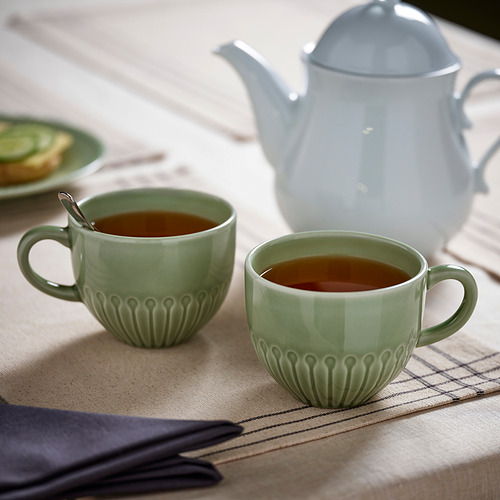 Dos tazas de té verde menta con asas están sobre un mantel beige junto a una tetera blanca. Las tazas están llenas de té.