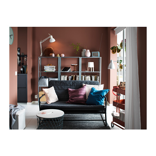 Habitación moderna con paredes de color rosa oscuro, sofá con estructura de metal negro, cojines coloridos, alfombra gris y estantes con una lámpara y libros.