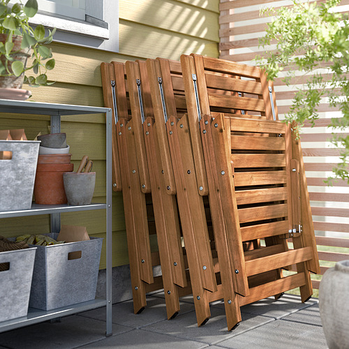 Sillas plegables de madera marrón apiladas al aire libre contra la casa, cerca de plantas en macetas y cestas de almacenamiento.