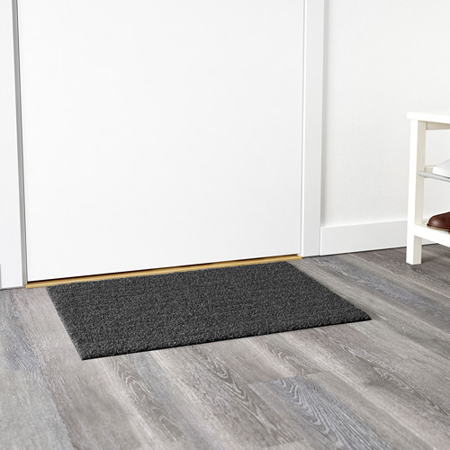 Felpudo rectangular de color gris oscuro colocado contra una pared blanca sobre un suelo de madera, diseñado para su uso en exteriores.