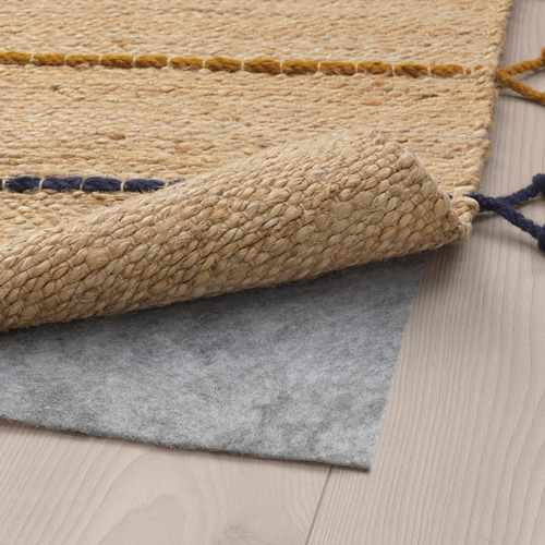 Una alfombra tejida de color beige con detalles en amarillo y azul descansa sobre un piso de madera, mostrando su parte inferior con respaldo de fieltro gris para evitar resbalones.