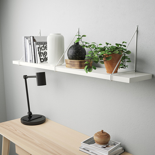Estante de pared blanco que sostiene libros, jarrones y una planta con una lámpara de escritorio ajustable negra junto a un escritorio de madera clara.