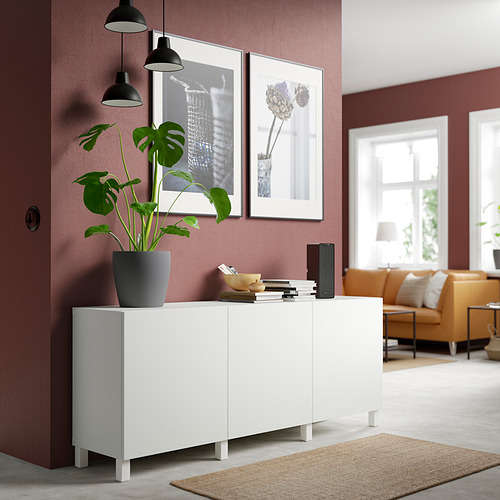 Salón moderno con aparador blanco BESTÅ. Sostiene planta, libros y altavoz. Entre paredes rojas, bajo lámparas colgantes, junto a un sofá naranja.