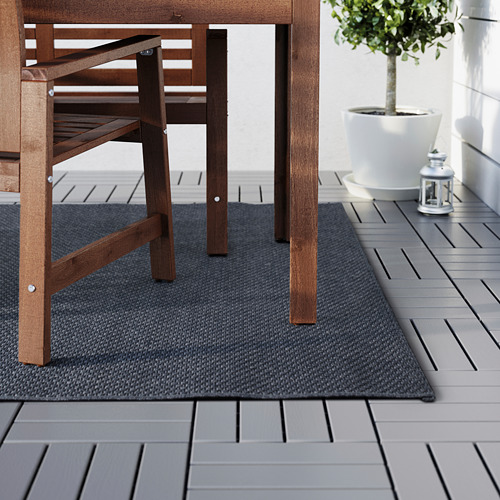 Una alfombra de exterior debajo de una mesa y sillas de madera, que protege las baldosas de la terraza y agrega comodidad. Una planta en maceta y una linterna decoran el lado.