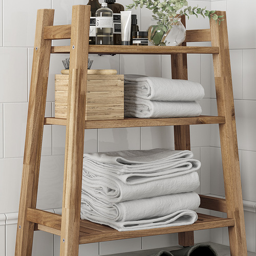 Estantería de baño de madera con tres niveles. La parte inferior contiene toallas dobladas, el medio tiene toallas y canasta, la parte superior exhibe artículos de tocador y plantas.