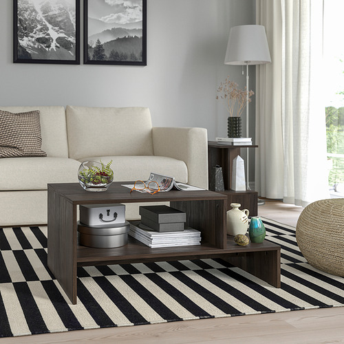 Mesa de centro HOLMERUD de color marrón oscuro con forma distintiva y almacenamiento abierto. Sobre alfombra de rayas, cerca de sofá beige y mesa auxiliar con lámpara.