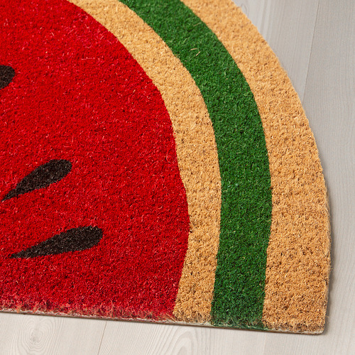 Felpudo rojo brillante con diseño de semillas negras, bordeado por rayas verdes y beige, colocado sobre suelo de madera clara.