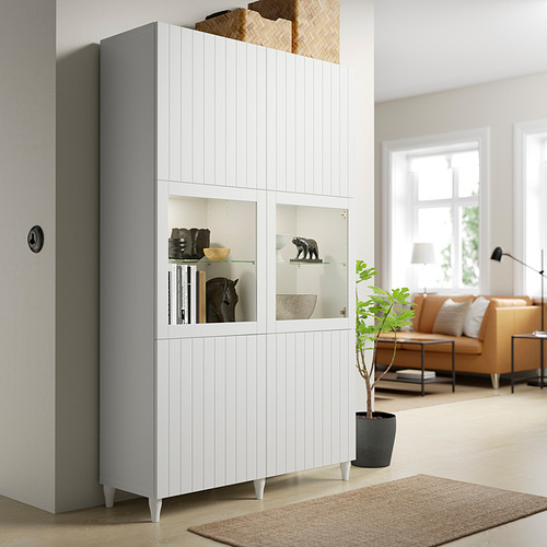 BESTÅ storage with glass doors, displaying items, beside a casting and plant, next to a living room with sofa and large window.