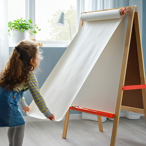 Dibujo infantil en rollo de papel grande con caballete de madera. Chica con vestido azul, cabello rizado, sosteniendo papel.