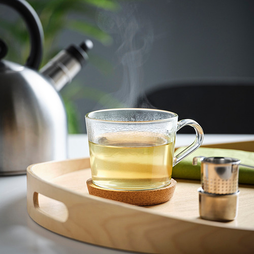 Una taza de vidrio en una bandeja de madera que contiene té humeante, colocada junto a una tetera y un colador.