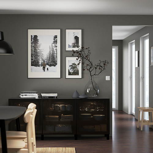 Comedor moderno con aparador BESTÅ negro, platos, ramas, libros, fotos de la naturaleza, lámpara colgante y sillas ligeras alrededor de una mesa negra.