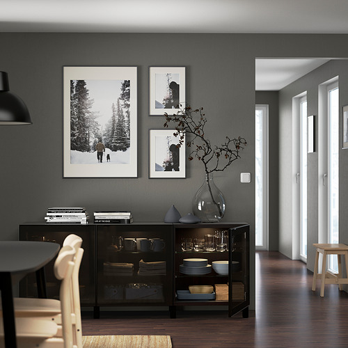 Sala de estar moderna con almacenamiento BESTÅ ajustable en la pared con puertas de vidrio que exhiben platos y un jarrón decorativo, junto con obras de arte enmarcadas.