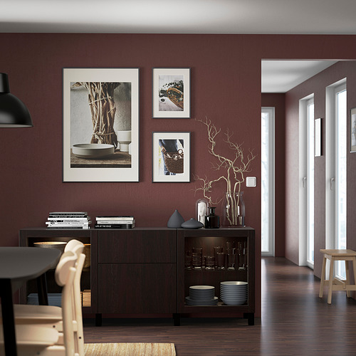 Aparador BESTÅ de madera oscura contra la pared burdeos con obras de arte. Es amplio, casi del suelo al techo, con platos apilables y jarrones decorativos.
