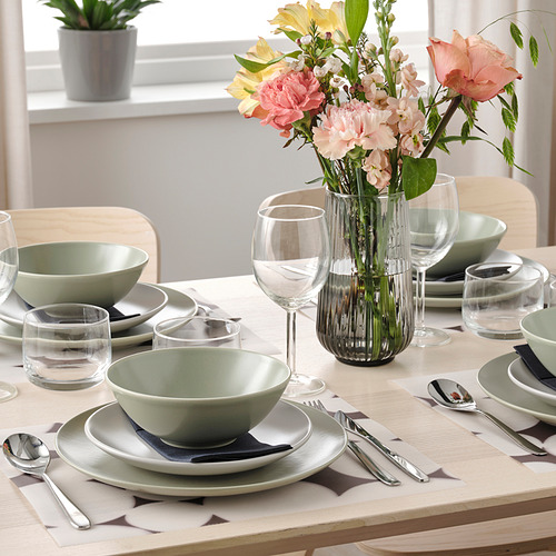 Mesa con platos verdes, vasos, cubiertos, manteles individuales y un jarrón con flores.