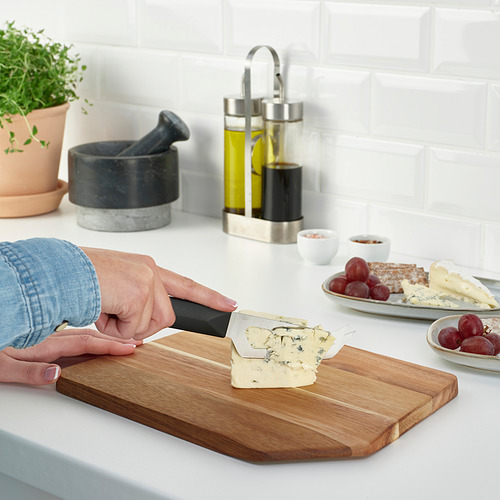 Persona cortando queso en una tabla de cortar de madera en una encimera de cocina. Cerca hay uvas, galletas saladas y condimentos.