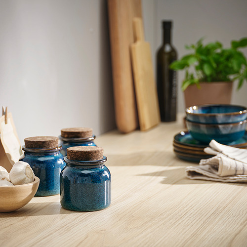 Tres tinajas de cerámica azul con tapas de corcho sobre mesa de madera, platos, cuencos apilados, botella y planta.