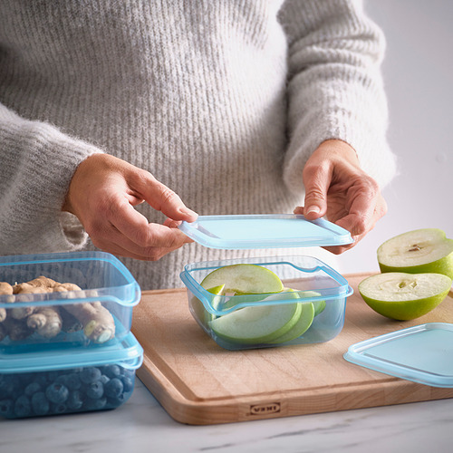 La persona coloca la tapa en un recipiente de comida azul con rodajas de manzana, mostrando un práctico almacenamiento de alimentos.