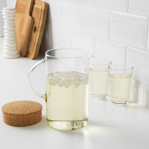 Jarra y tazas de vidrio transparente colocadas sobre una encimera blanca, llenas de una bebida ligera y cubitos de hielo, acompañadas de una tabla de madera y posavasos.