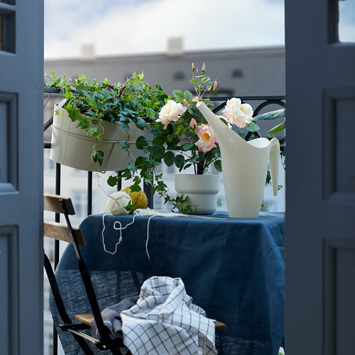 Escena del balcón: macetas de madera clara con flores rosas, regadera blanca, mantel azul, silla de metal negro con una manta a cuadros.