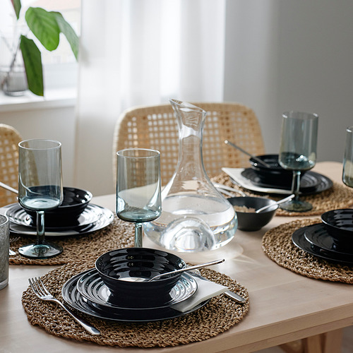 Mesa de comedor con cristalería y platos negros sobre manteles individuales tejidos, con un diseño apilable, apto para lavavajillas y elegante.