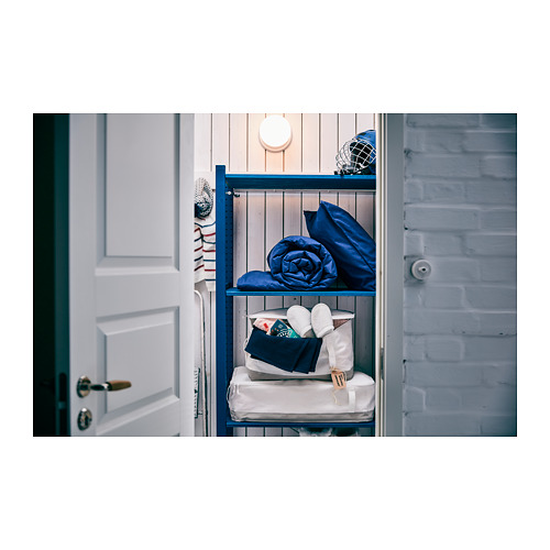 Mueble de almacenaje de madera azul contra una pared de ladrillo claro, con ropa de cama blanca y azul y una lámpara en el estante superior.