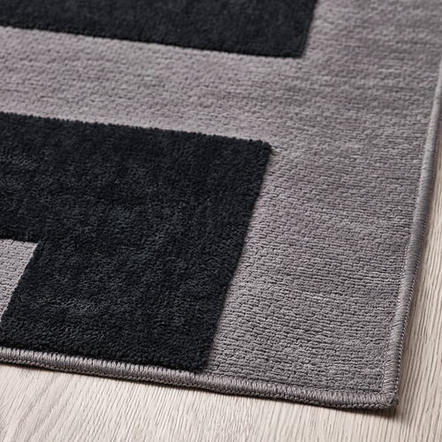 Alfombra gris con secciones negras dispuestas en un patrón geométrico moderno. Suave, con una superficie texturizada, colocado sobre pisos de madera noble.